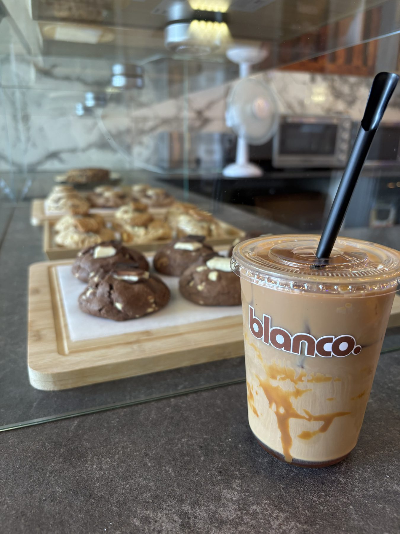 An iced Blanco latte on the counter, with cookie trays in the pastry case behind it.
