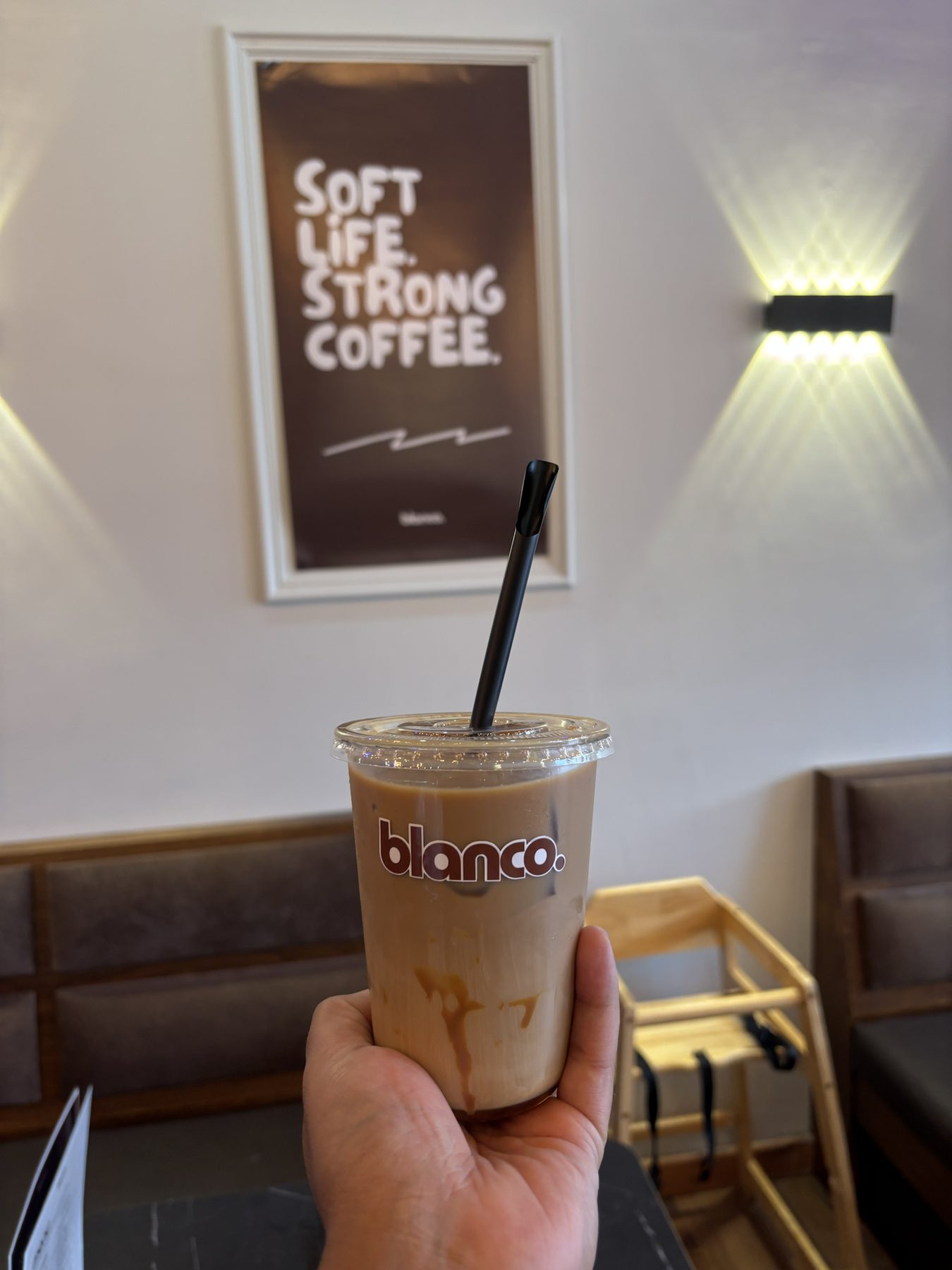 An iced Blanco latte held in front of the Soft Life Strong Coffee print.